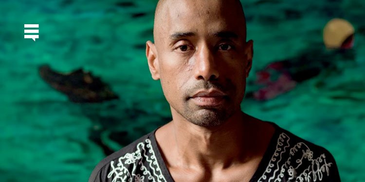 O artista Antonio Obá, homem negro, careca, usando vestes pretas. Seu semblante é sério.