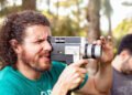 Homem segurando uma câmera filmadora Super 8mm, ao ar livre. Ele veste uma camiseta verde e sua feição é concentrada.