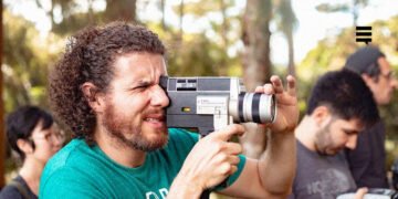 Homem segurando uma câmera filmadora Super 8mm, ao ar livre. Ele veste uma camiseta verde e sua feição é concentrada.