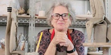 Mulher branca, cabelo grisalho curto, apoiada em uma bancada, com as mãos unidas. Ela usa um avental cinza, blusa violeta estampada com formas geométricas e óculos de grau. Seu semblante é alegre.