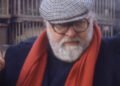 Homem branco, cabelo e barba grisalha, usando casaco azul, cachecol vermelho e boina xadrez. Seu semblante é sério e ele está com o dedo da mão direita apontado para o alto.