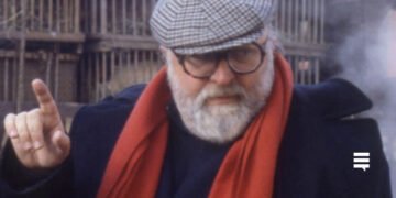 Homem branco, cabelo e barba grisalha, usando casaco azul, cachecol vermelho e boina xadrez. Seu semblante é sério e ele está com o dedo da mão direita apontado para o alto.