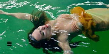 Mulher branca usando vestido amarelo, cabelo comprido castanho escuro, jogada em uma piscina com fundo verde.