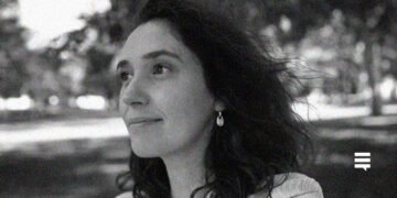 Foto preto e branco de uma mulher branca, de cabelo comprido até os ombros, de perfil. Ela usa um brinco na orelha esquerda. Seu semblante é alegre. Ela está ao ar livre.