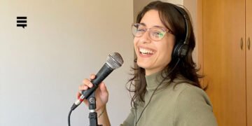 Mulher branca, cabelo comprido castanho, usando óculos de grau e camisa de gola alta verde musgo. Ela segura um microfone em um pedestal. Seu semblante é alegre, ela sorri com a boca aberta.