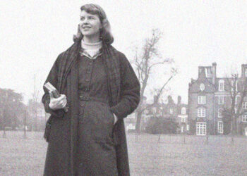 Foto preto e branco da poeta Sylvia Plath segurando livros e com a mão no bolso de seu vestido. Ela também usa um capote. Seu semblante é alegre. Ela está ao ar livre e não olha para a foto.
