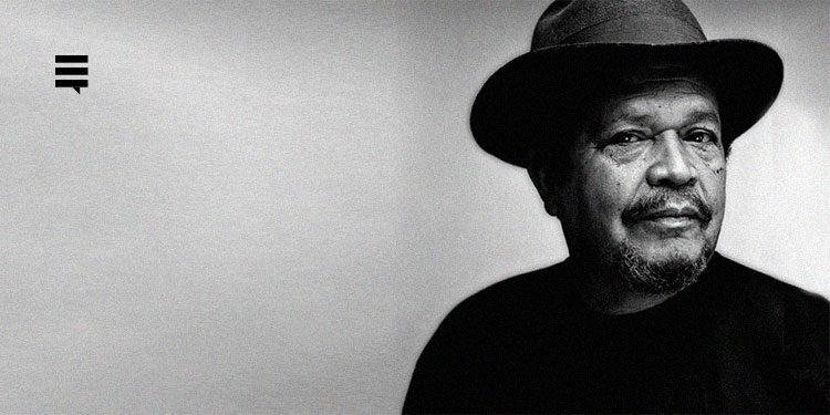 Walter Firmo em fotografia preta e branca. Ele é um homem negro, usando camisa preta e chapéu, com o rosto de perfil voltado para a direita. Ao fundo, uma parede.