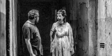 Foto preto e branco de homem branco, usando camiseta escura, cabelo curto e barba, ao lado de uma mulher branca, usando vestido claro. Eles conversam e o semblante deles é sério.