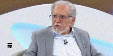 Homem branco, cabelos e barba grisalhos, usando óculos de grau, camisa social azul clara e blazer cinza, sentado em uma cadeira dentro de um estúdio de televisão