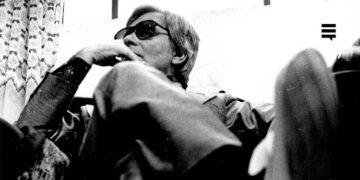 Foto preto e branco de homem branco, sentado, com as pernas cruzadas, usando óculos escuro. Seu semblante é sério. Cabelo curto.