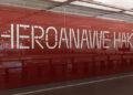 Foto de uma parede vermelha com o nome do artista Sheroanawe Hakihiiwe escrito em branco